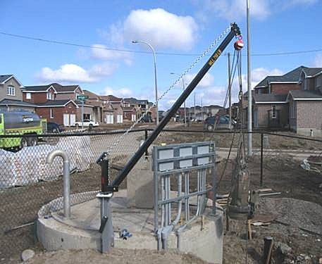 A long-radius eme davit crane mounted in a circular concrete utility vault at a residential subdivision under construction, the arm extended out over the access opening.