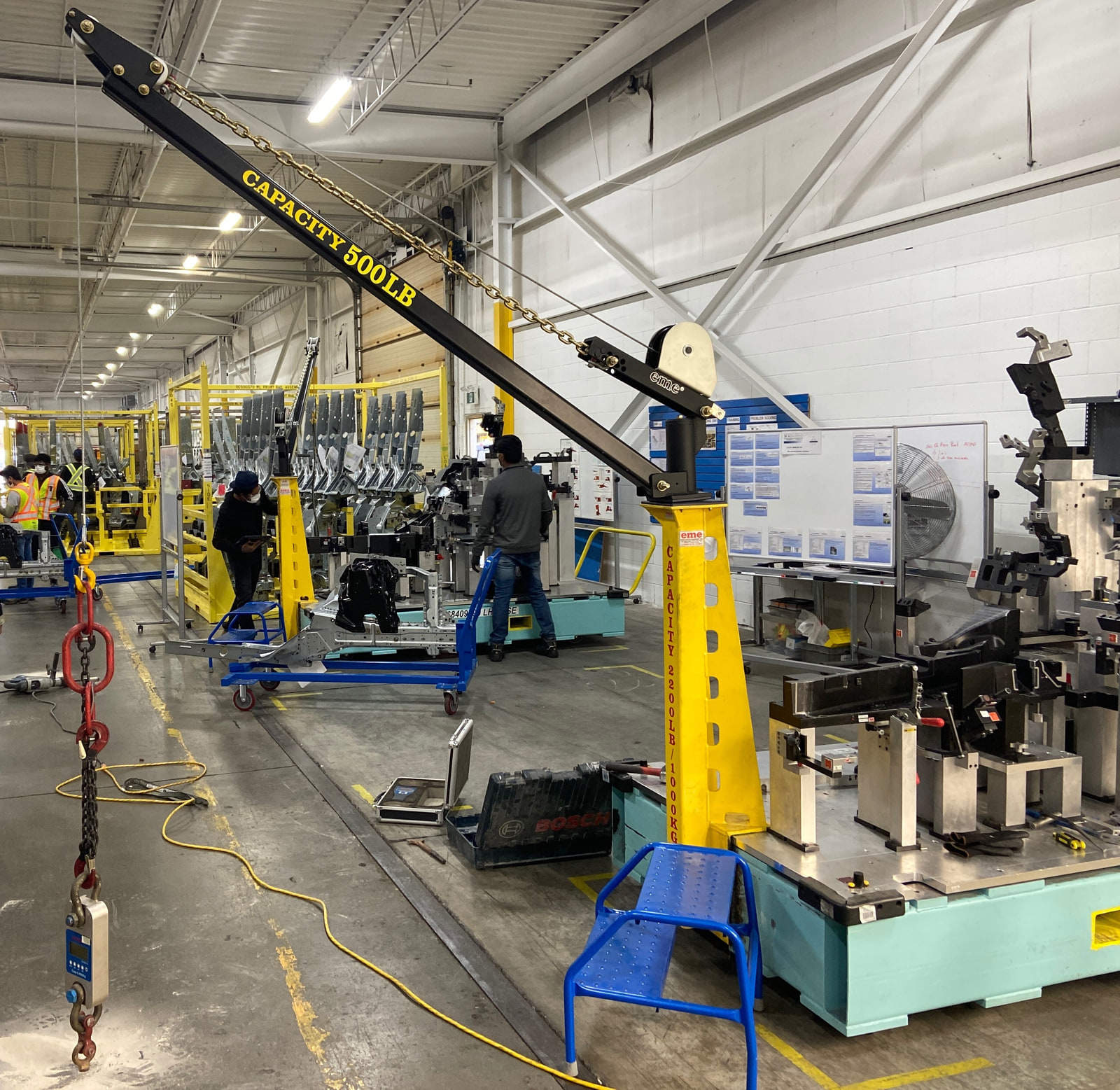 A safety-yellow eme davit crane rated 500 lb standing on the floor of a manufacturing plant among assembly-line equipment, positioned for fixture and tool-change lifts.