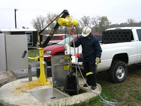 An eme davit crane in a safety-yellow rim socket beside a wastewater wet well, a hard-hatted worker raising a submersible pump on the hoist chain, a service truck parked behind.