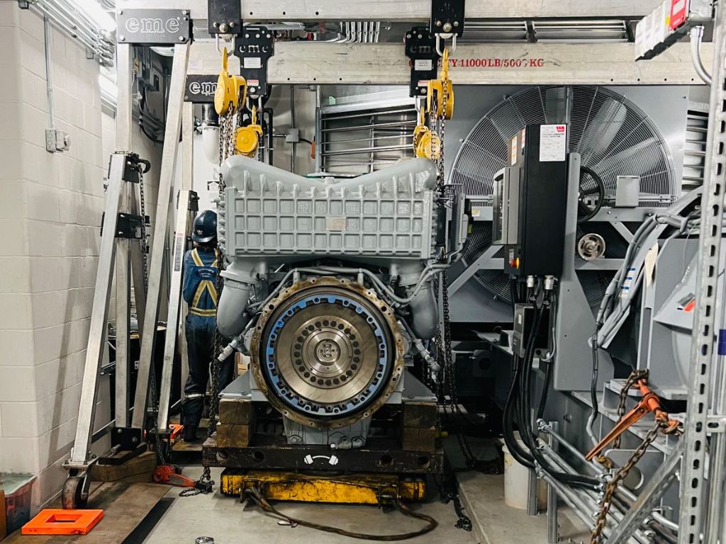 Two eme 11000 lb capacity aluminum gantry cranes in a paired-lift configuration suspending a large industrial engine block during a maintenance operation; large cooling fans and equipment racks visible in the background.