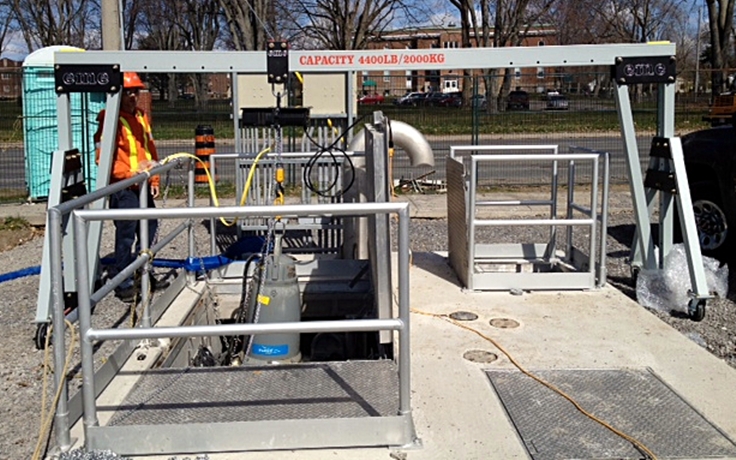 A 4400R aluminum gantry crane outdoors over a wastewater pump station, pulling a submersible pump while a worker in an orange safety vest positions the lift.