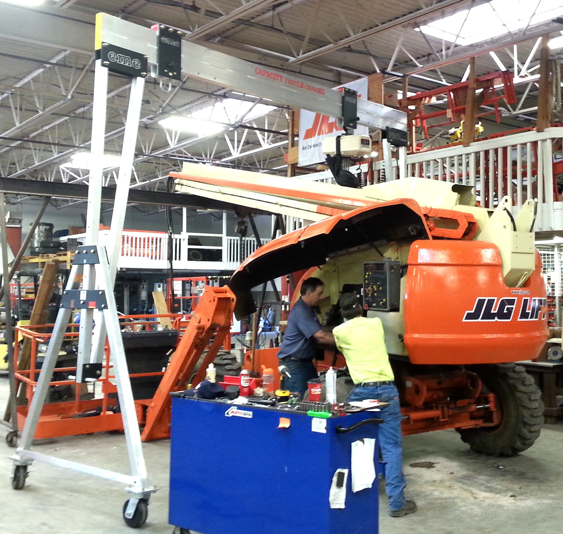 An eme 6600 lb capacity aluminum gantry crane positioned above an orange JLG aerial work platform inside an equipment-rental maintenance facility, with two technicians servicing the boom lift's powertrain.